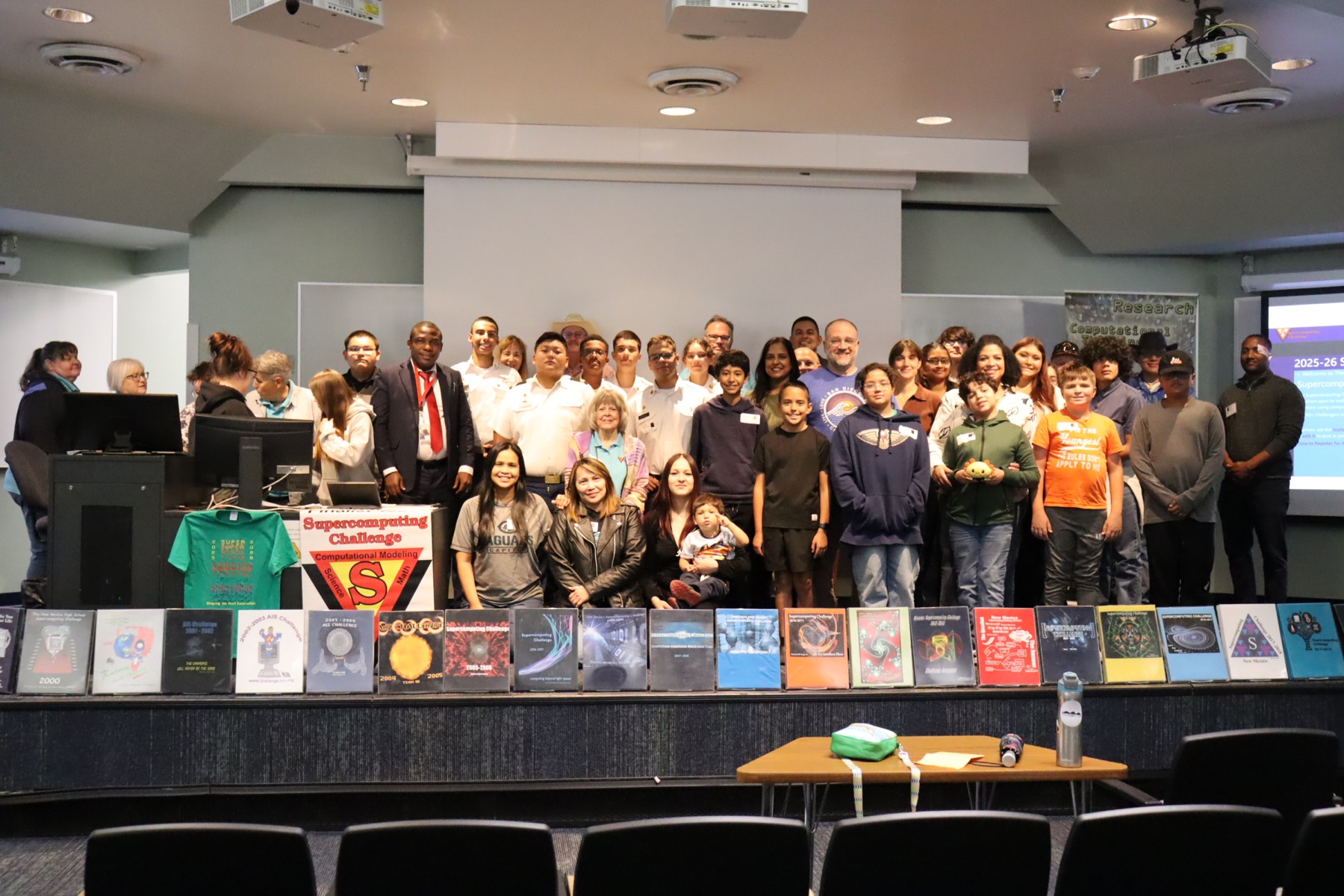 Students, teachers, and mentors posing on stage at Kickoff, with Challenge yearbooks displayed in front
